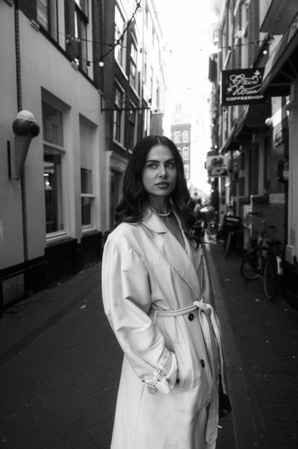 a black and white photo of a woman in a trench coat in front of a coffee shop