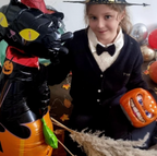Young girl in witch hat with inflatable black cat and pumpkin toy. Fall leaves on dark floor create a festive, Halloween mood.