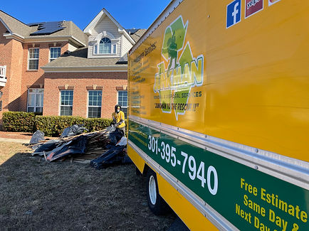 A Junkman Removal & Disposal Services truck parked in front of a residential property with a worker managing a pile of construction debris and black trash bags. The branded truck features the company's contact information and services such as free estimates, same-day service, and next-day service availability. The image highlights a professional junk removal service in action, focusing on residential debris cleanup and eco-friendly disposal solutions