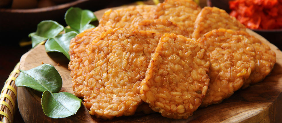 Golden brown tempeh slices on a wooden board with green leaves, set against a blurred background with red hues, creating a warm, rustic mood.