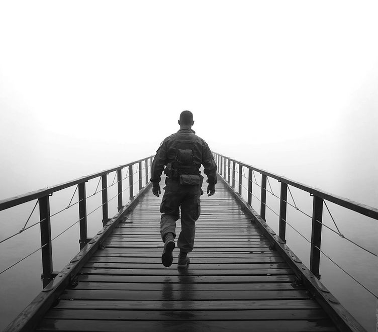 A solitary soldier, seen from behind, walks across a long, wet wooden bridge on a foggy da