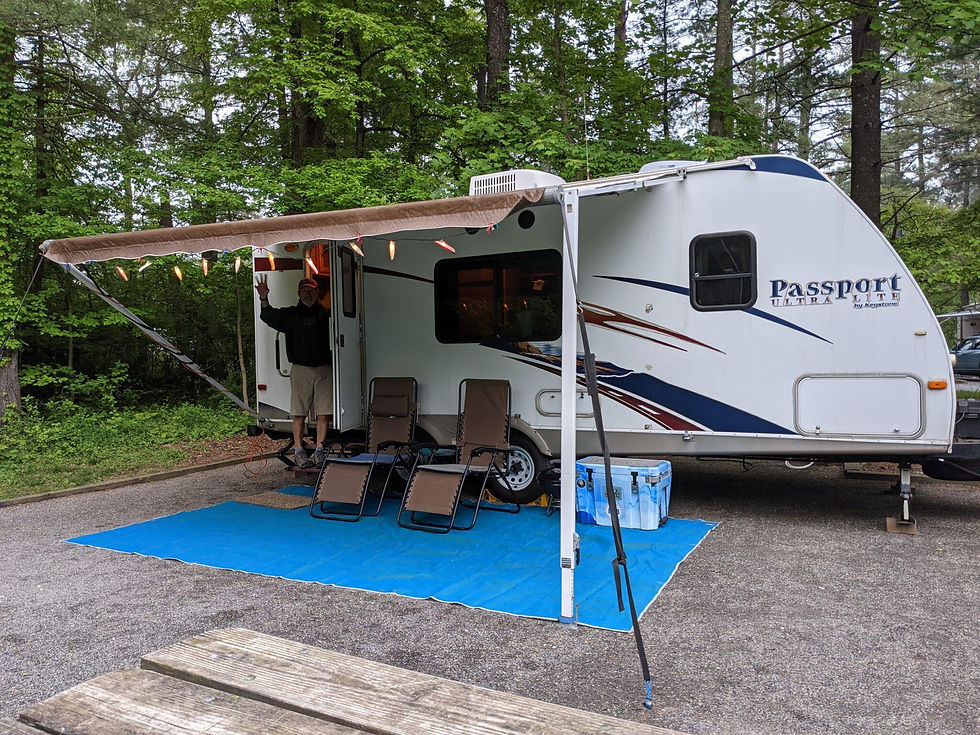 Man waving from RV door in forest camp, chairs on blue mat under awning. "Passport Ultra Lite" text on RV, festive lights hanging.