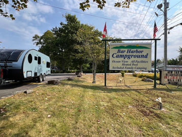 Blue trailer and pickup on road by "Bar Harbor Campground" sign. Trees and blue sky in the background. Flags waving gently.