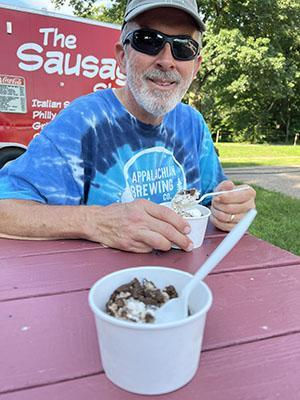 Man in sunglasses, tie-dye shirt smiles holding ice cream at a red picnic table. Sausage shop truck in background. Sunny day.