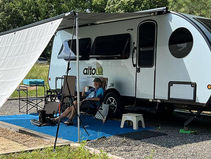 Man relaxes on a chair with a dog beside him outside a white camper under a canopy. Trees in the background, "alto" logo on camper.