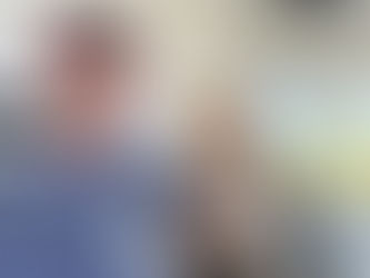 Man in a cap smiles while holding a drink outdoors. He's seated near wooden barrels under a white porch. Blue sky in the background.