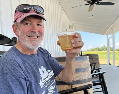 Man in a cap smiles while holding a drink outdoors. He's seated near wooden barrels under a white porch. Blue sky in the background.