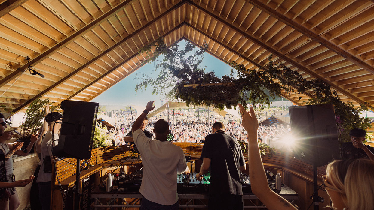 Photo of Dr Dubplate & BlueTooth playing Lost Gardens stage captured from behind, so we see them behind the decks with a crowd of people beyond the enclosed stage roof