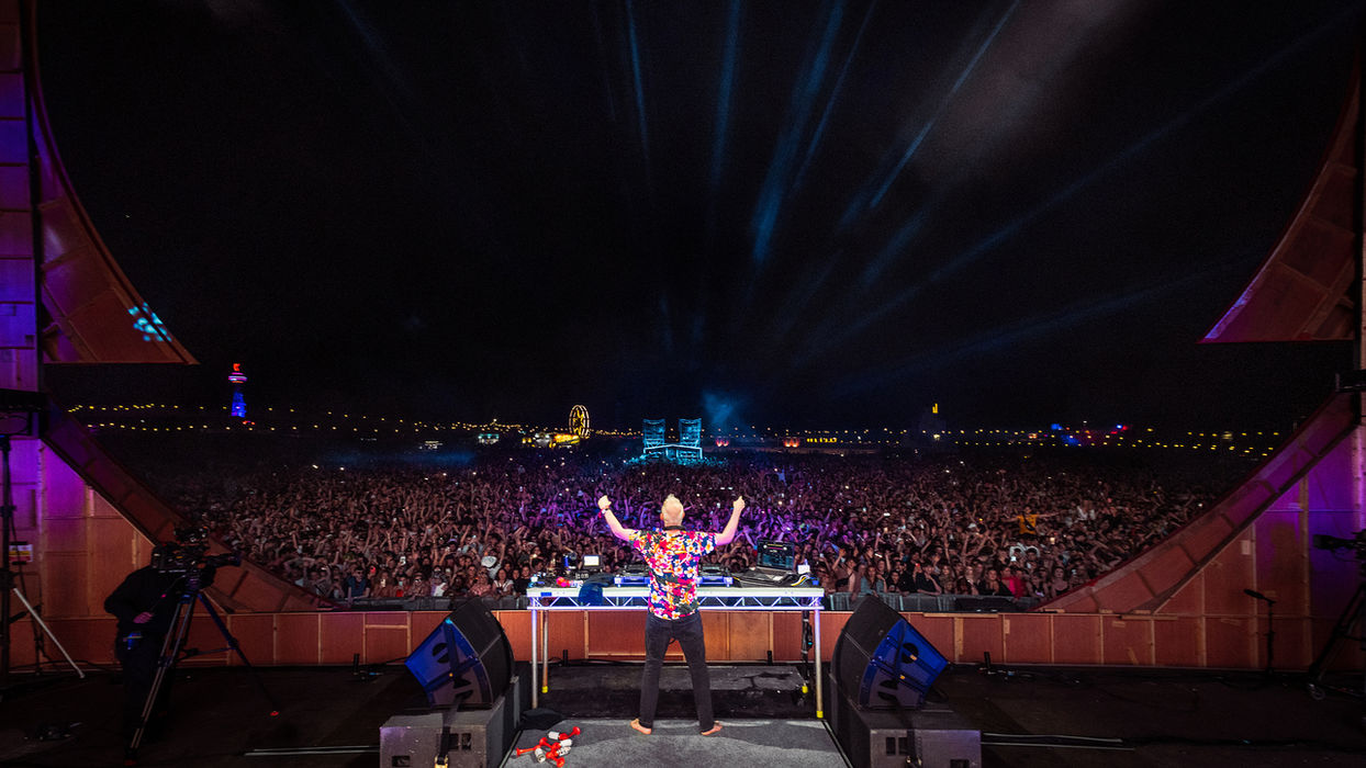 Fatboy Slim stands behinds the decks of the Love Saves stage, we see the bare side of the frontage and stage as this shot is from behind, and the huge crowd lit by the stage projections in the dark night