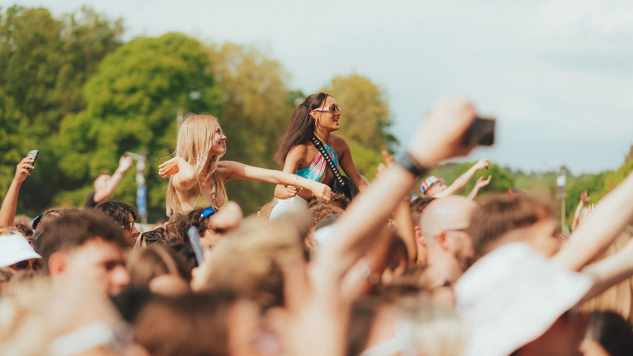 Two femme-presenting individuals sat on the shoulders of unseen people in the middle of a crowd with their arms in the air.