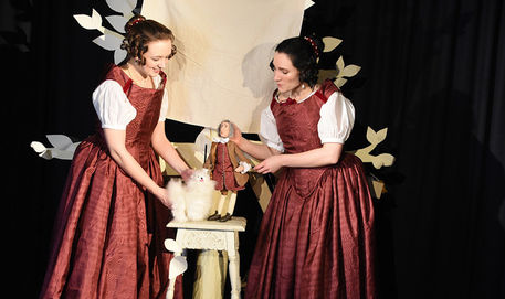 Katie to the left with Eleanor to the right, both wearing red 17th century gowns. Katie is puppeteering Diamond, a small, fluffy, white dog while Eleanor puppeteers Sir Isaac Newton, who looks rather austere!