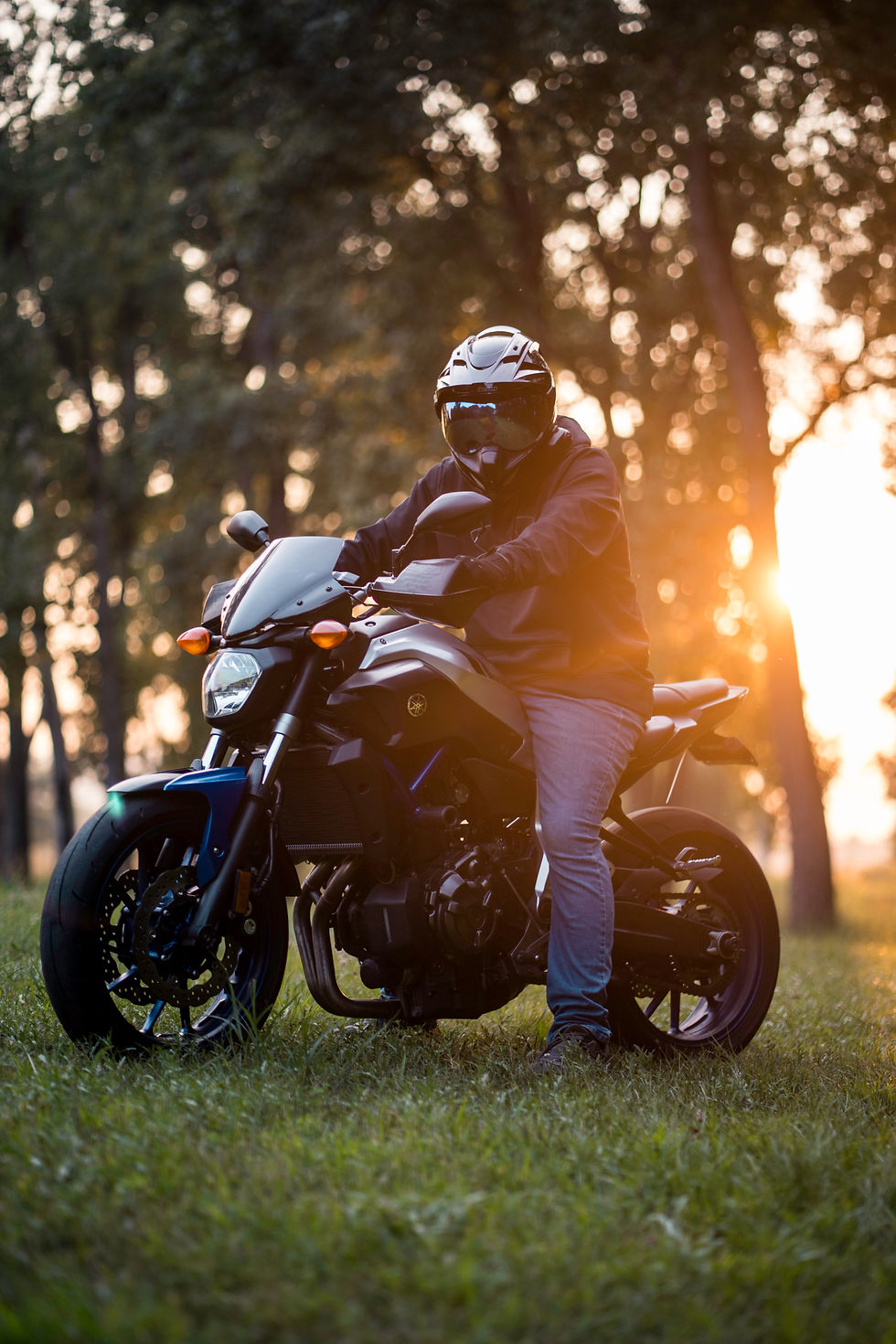 A person in a helmet and dark clothing sitting on a Yamaha motorcycle in a grassy field at golden hour, with warm sunlight filtering through trees in the background.