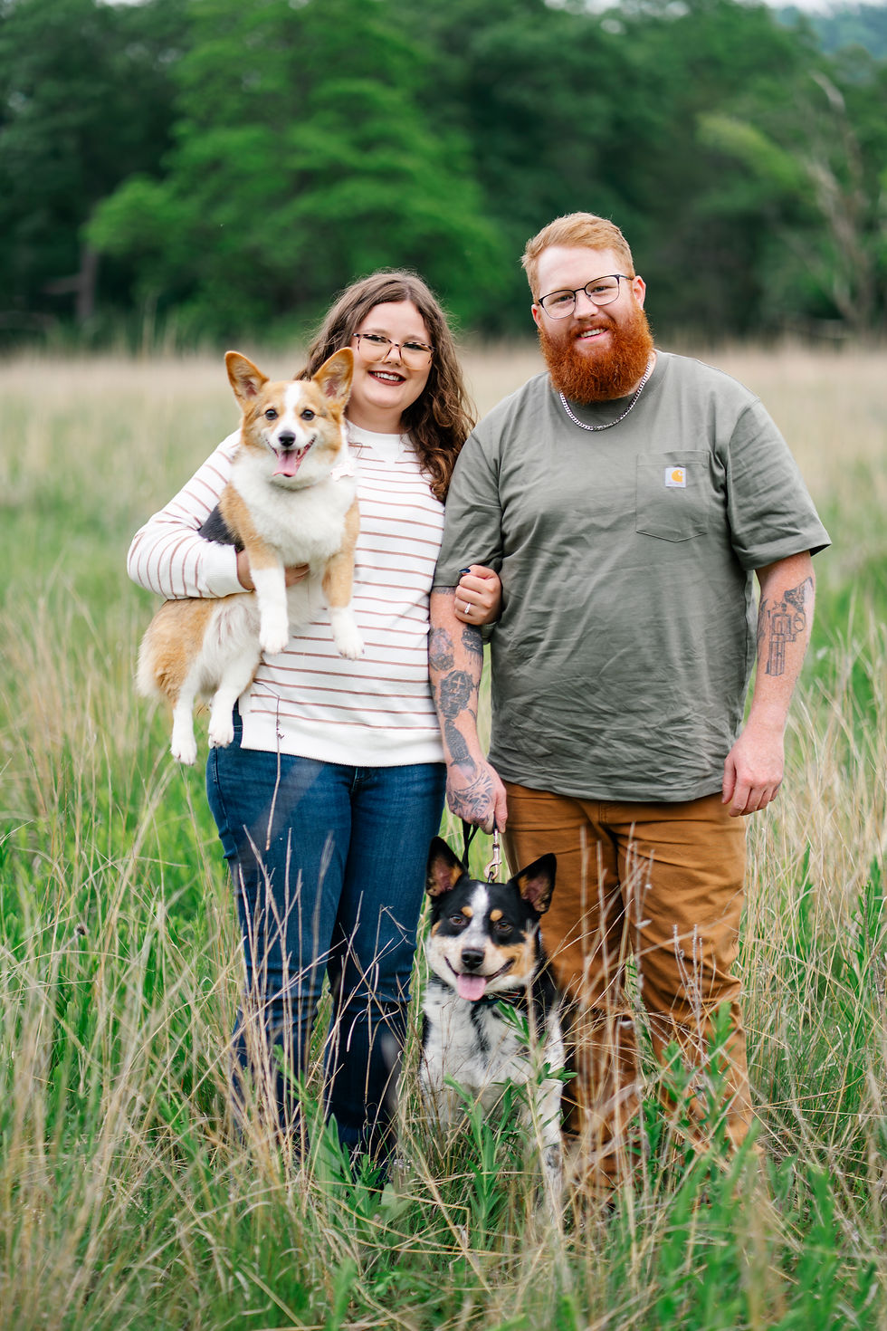 Couple standing in a tall grassy field with two happy dogs, one being held and one on a leash, smiling during their engagement photo session