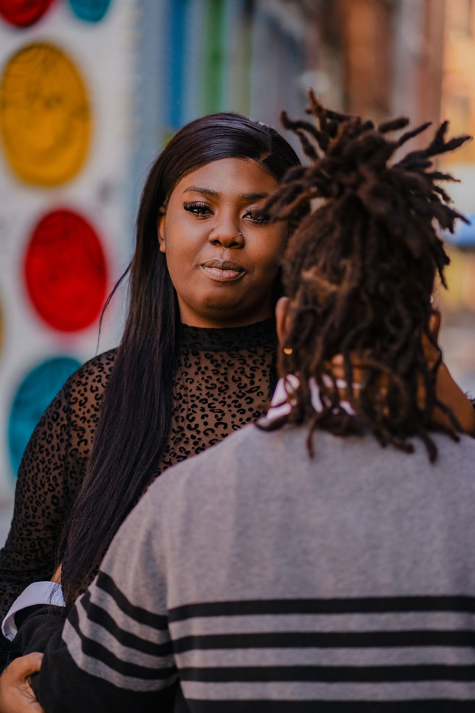 A Black couple stands close together in front of a colorful mural. One person faces the camera confidently while the other gently holds their back, partially out of frame.