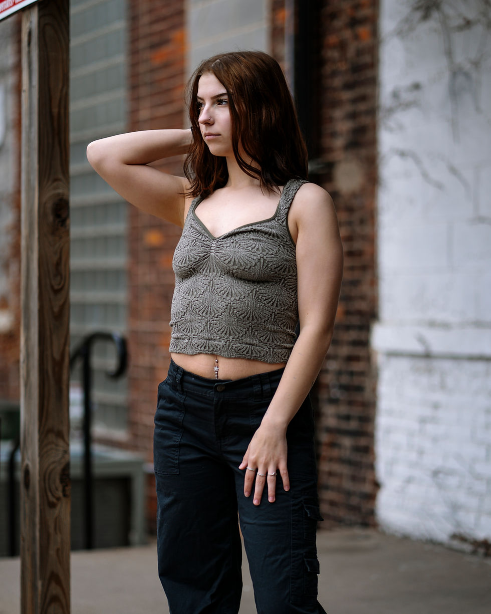 Woman in a patterned tank top and black pants stands against a brick wall, hand on head, looking thoughtful. Urban setting, muted tones.