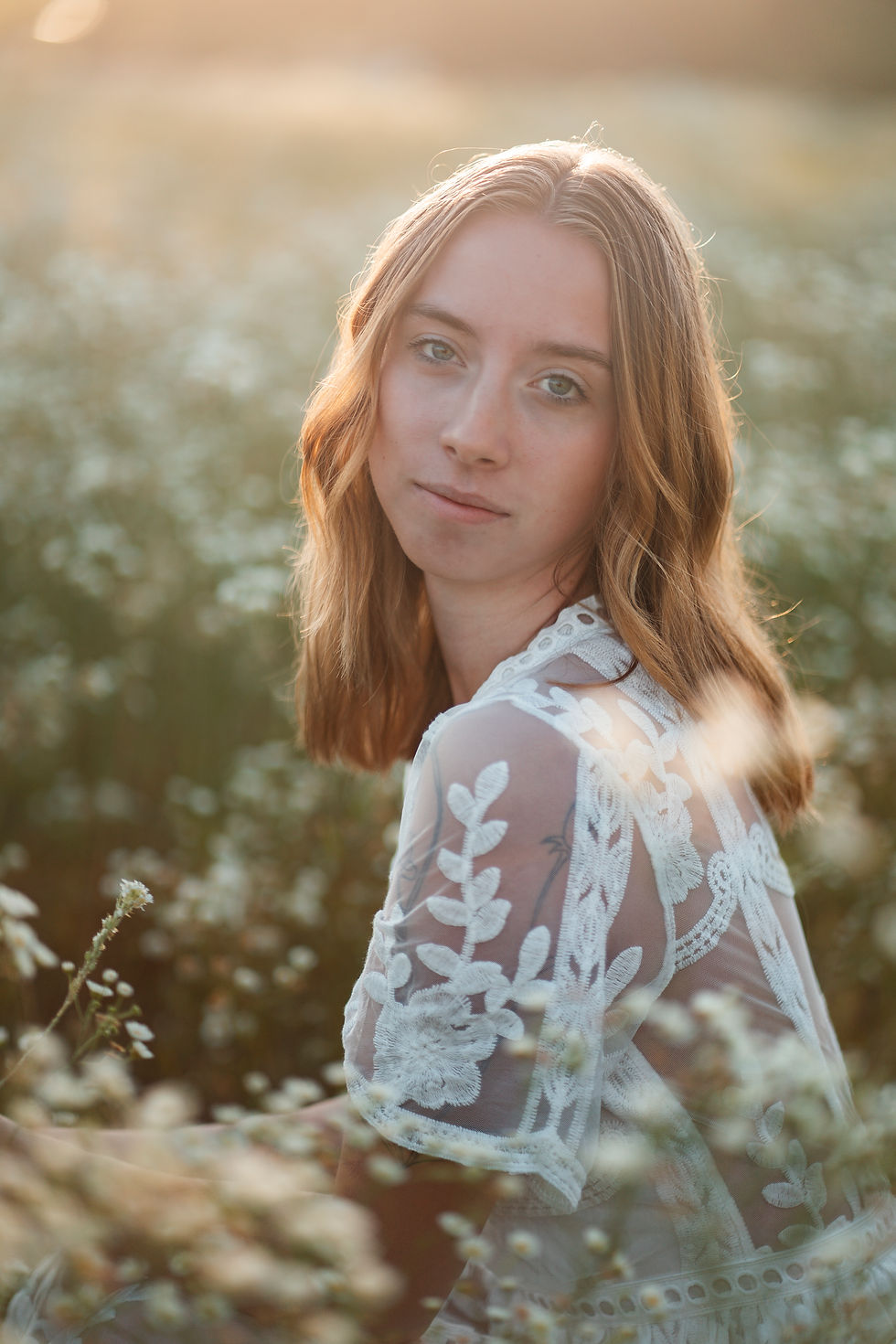 A young woman with shoulder-length blonde hair in a sheer embroidered dress sitting in a field of wildflowers, softly lit by golden morning sunlight.