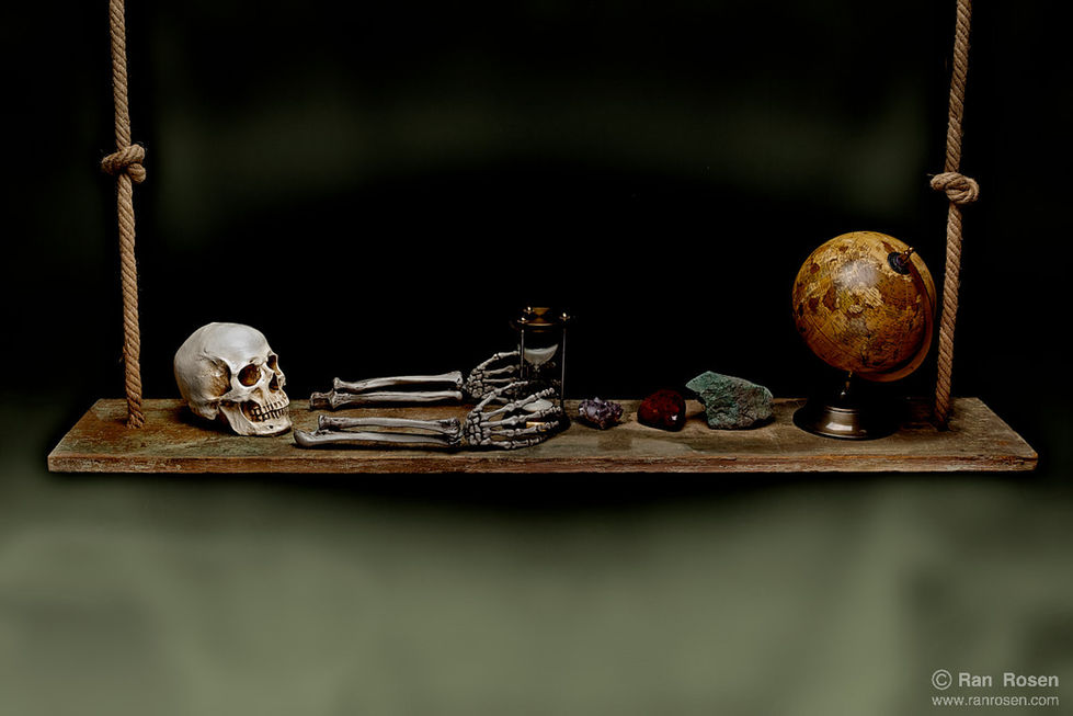 A still-life arrangement on a suspended wooden plank: a human skull and skeletal hands on the left, an hourglass in the center, followed by stones and minerals, and a small aged globe on the right, all set against a dark background.