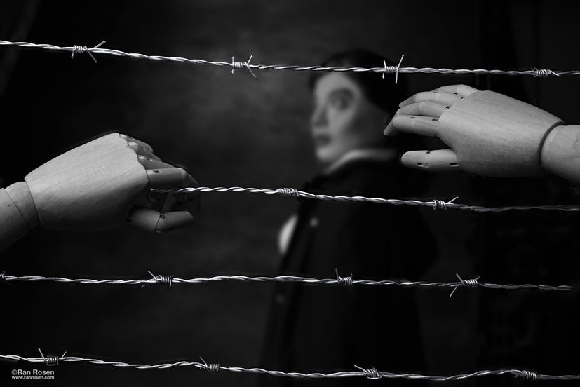 Two hands reaching toward each other through barbed wire, with a blurred figure in the background; symbolic still-life photograph by Ran Rosen depicting loss and separation after October 7.