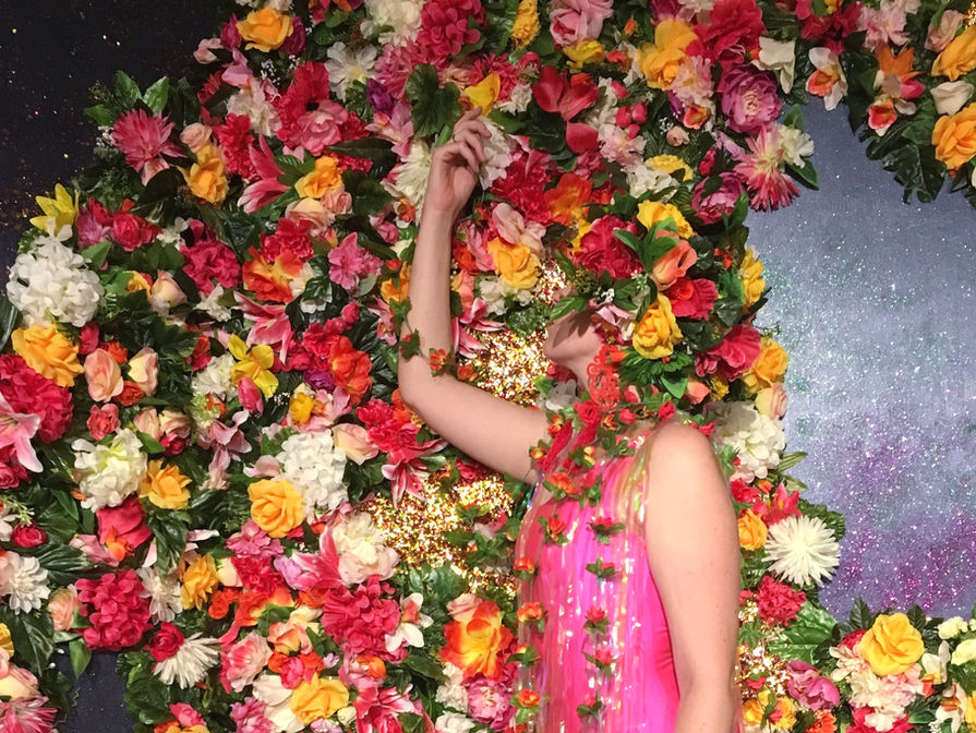 Landscape image of a multi-coloured floral backdrop with a section of confetti leading into a glittery navy section in the centre right, and top left. A white woman stands sideways facing towards the left of the frame in front of the wall wearing a pink leotard and floral wreaths. Her right arm is raised in front of her face and she is looking at it. Her head and  parts of her body are disguised by flowers and vines, blending into the floral wall. 