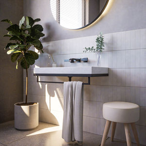 A sleek, modern concrete basin (Penny Sink) with sharp geometric detailing, mounted on a minimalist black metal frame, set against a tiled wall with natural light softly illuminating the scene. Complementary decor includes a round backlit mirror, a potted plant, and a stool with wooden legs.