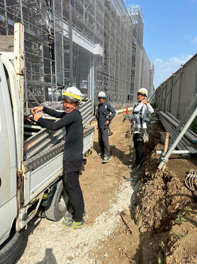 建設現場で作業する三人の作業員。資材を運搬中。背景には足場、株式会社 渉工業。