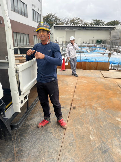 建設現場の作業員が笑顔で立つ、トラックの横。背景に建物と求人情報。株式会社 渉工業。