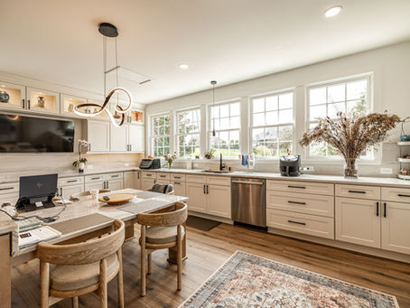 Bright, spacious kitchen with white cabinetry, a long counter beneath several large windows, stainless steel appliances, a central island with woven chairs, pendant lighting, and a patterned area rug.