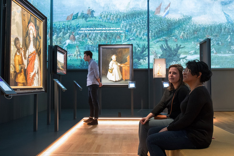 Three people in a room with brightly lit paintings mounted on posts, and a large image of a battle scene projected on the wall