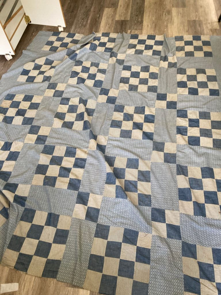 Blue and white checkered quilt on a wooden floor. The quilt has striped patterns among plain squares, creating a cozy and rustic feel. This will be used in an Upcycle Sewing project
