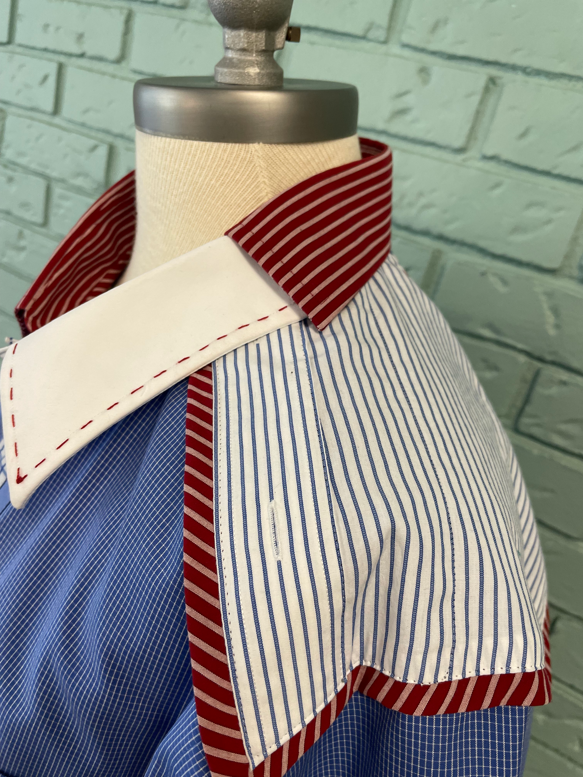A mannequin displaying a USA Collection shirt with a blue base, featuring a red and white striped collar, a white inner collar, and blue and white vertical striped accents on the yoke. The fabric edges are finished with red stitching, set against a textured, light green brick wall background.