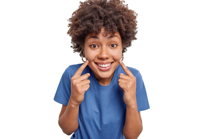 portrait-cheerful-curly-haired-young-woman-points-index-fingers-cheeks-smiles-broadly-feels-very-happy-dressed-casual-blue-t-shirt-isolated-white-background-look-my-glad-face (1).png