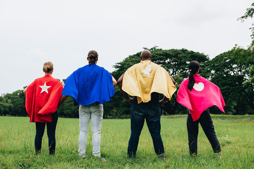 Four people stand heroically in a field wearing colorful capes with symbols—star, crescent, and heart—against a backdrop of green trees.