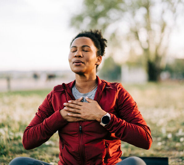 A person meditates in a grassy park, eyes closed, hands on chest. Wearing a red jacket and smartwatch, serene expression, blurred trees behind.
