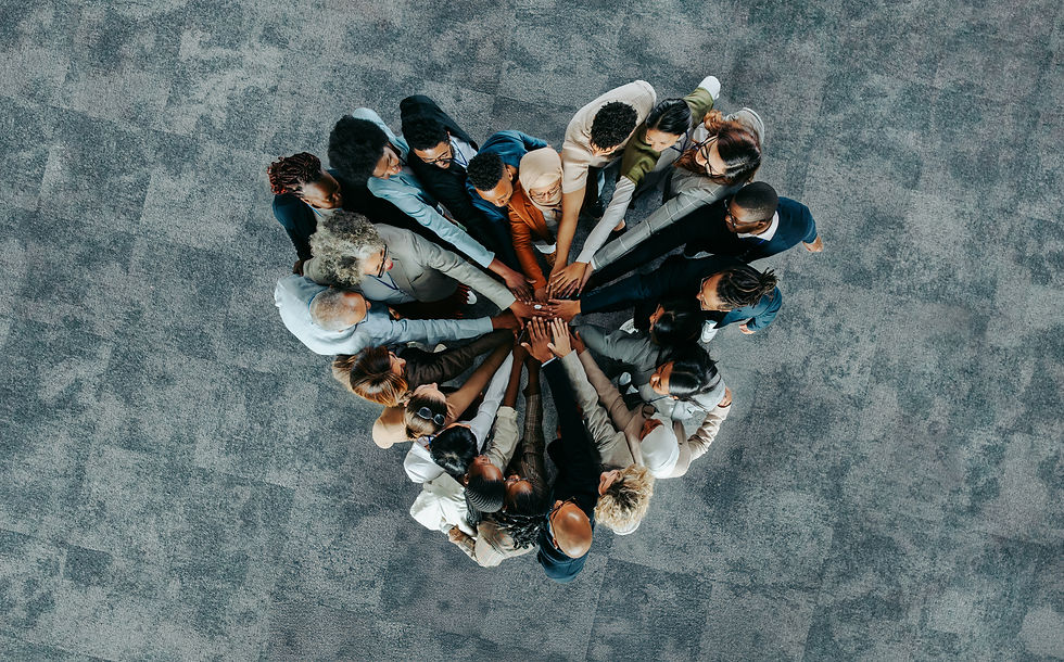 Diverse group of people in a circle place hands together in unity on a gray textured floor, symbolizing teamwork and collaboration.