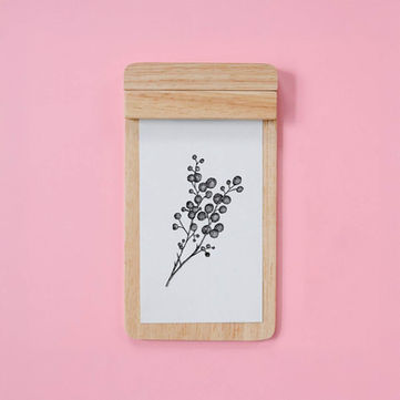 Black and white sketch of wattle on a white piece of paper in a wooden clipboard, on a pink background.