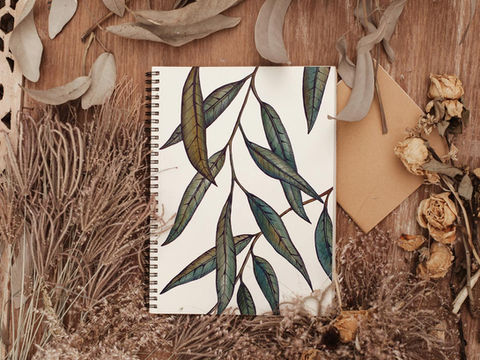 Watercolour painting of eucalyptus leaves in an art book, on a wooden desk amongst dried leaves.
