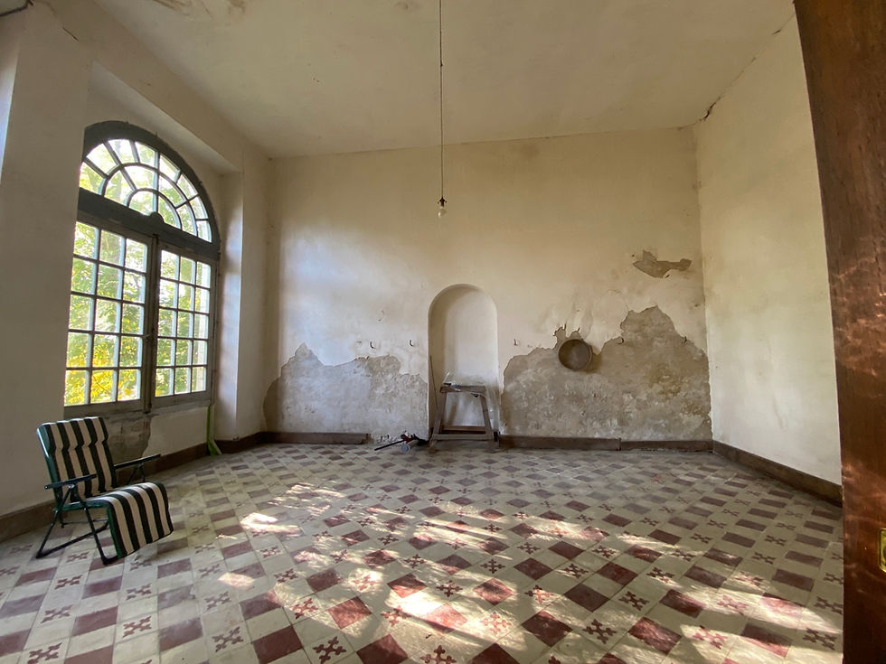 Old room with peeling walls, large arched window, patterned tile floor, and two striped chairs. Sunlight filters in, creating a calm mood.