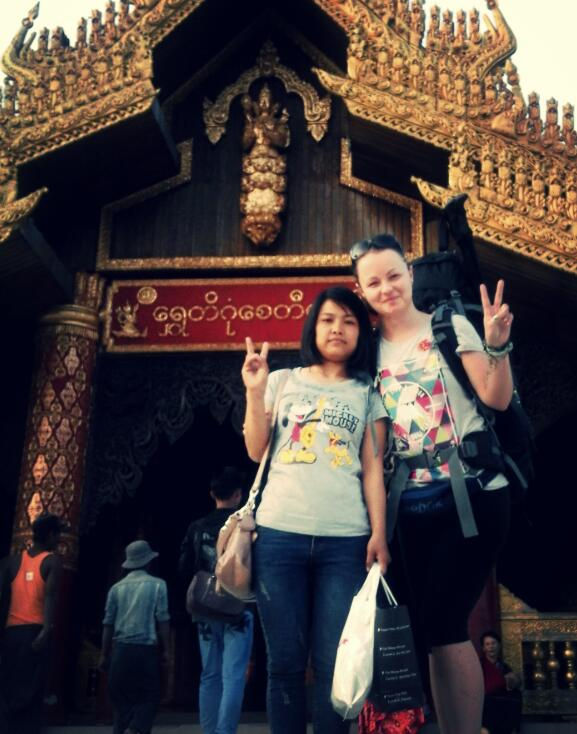 Two women pose with peace signs in front of an ornate, gold-decorated temple entrance. One holds shopping bags. Cheerful mood. Grumpy Nomad Cat