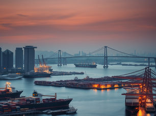 A cinematic editorial image of Singapore’s Marina Bay skyline and port at dusk, with massive shipping containers and cargo ships in the foreground. In the distance, fragmented trade routes represented by separate industrial structures are connected by elegant bridges converging on Singapore. Subtle visual contrast between walls/barriers and open bridges, conveying global trade fragmentation versus Singapore’s role as a connector. Realistic, sophisticated, corporate, no text, no people.
