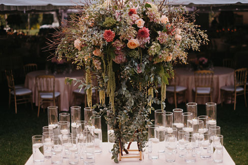 M&B's wedding day turned out to be an stunning celebration that guests will remember for years to come! The bride looked nothing short of radiant, wearing a beautiful gown that sparkled in shades of cream, ivory and gold. The ceremony was artfully decorated with wild garden flowers in soft pinks, vibrant peaches, and rich dark oranges, all surrounded by lush eucalyptus leaves, which together created a romantic and enchanting atmosphere. The reception came alive with the warm glow of candlelight where tables were adorned with vibrant pink, purple, and blue flowers cascading elegantly on the tables. This exquisite setup was further enhanced by the crystal and gold charger plates, complemented by lavender napkins and luxurious pink velvet linens, creating a breathtaking and unforgettable dining experience for everyone present.