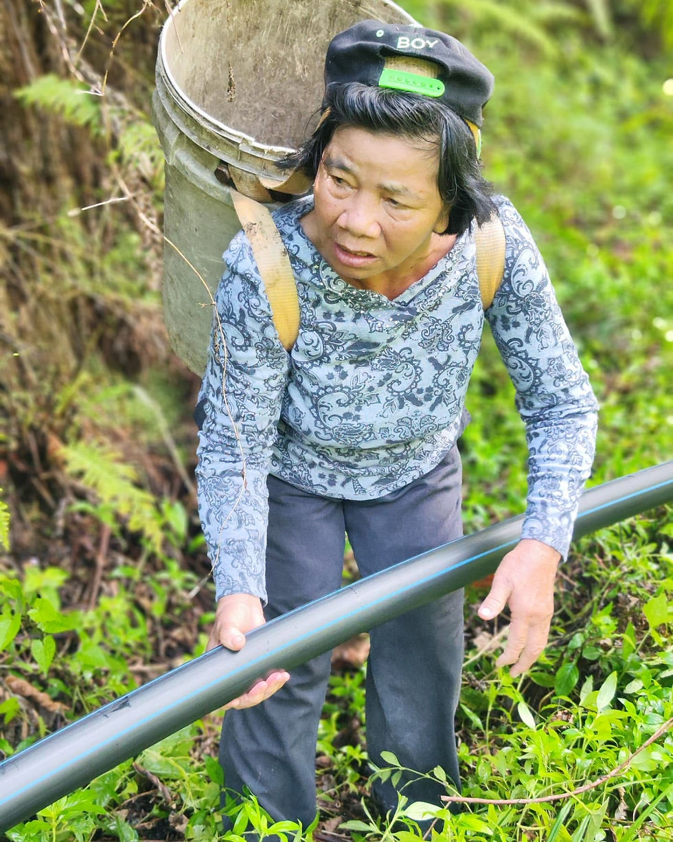Local villager holding and positioning a black HDPE water pipe with a blue stripe on a grassy hillside during a gravity-fed water supply programme in rural Sabah.