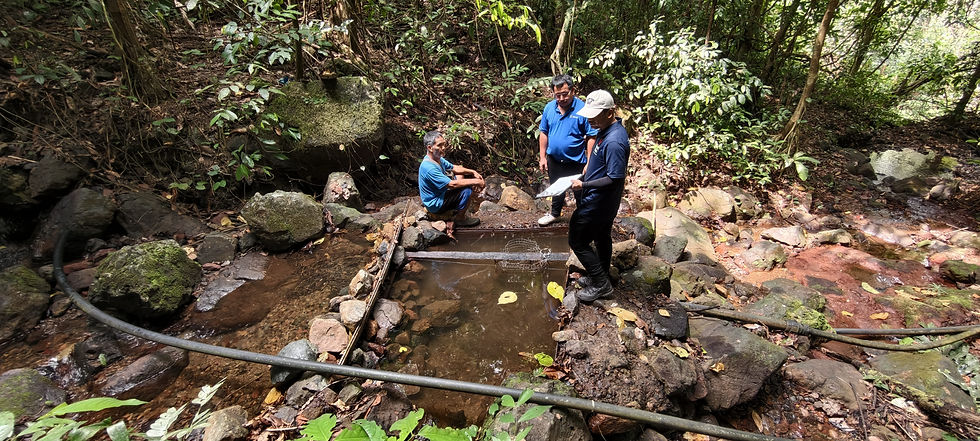 RuralKita field team member and villagers inspecting an existing water catchment structure and connected HDPE pipelines beside a rocky forest stream in Kampung Talukung, Kota Belud, Sabah.
