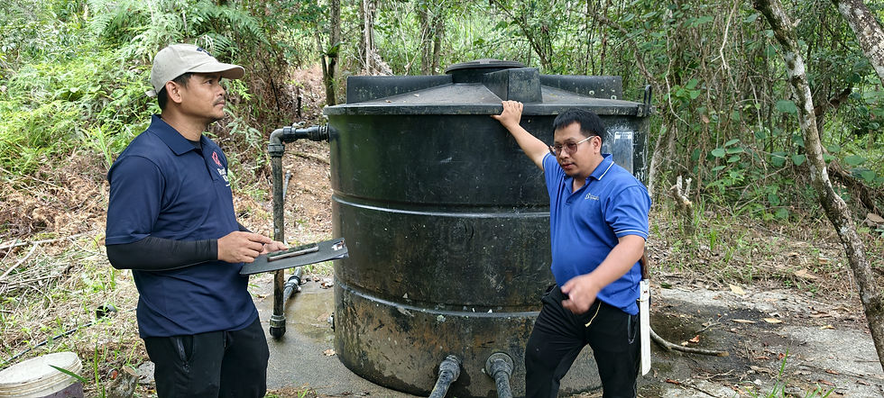 RuralKita team inspecting a large black water storage tank with a villager and its connected pipelines in a forested area of Kampung Talukung, Kota Belud, Sabah.
