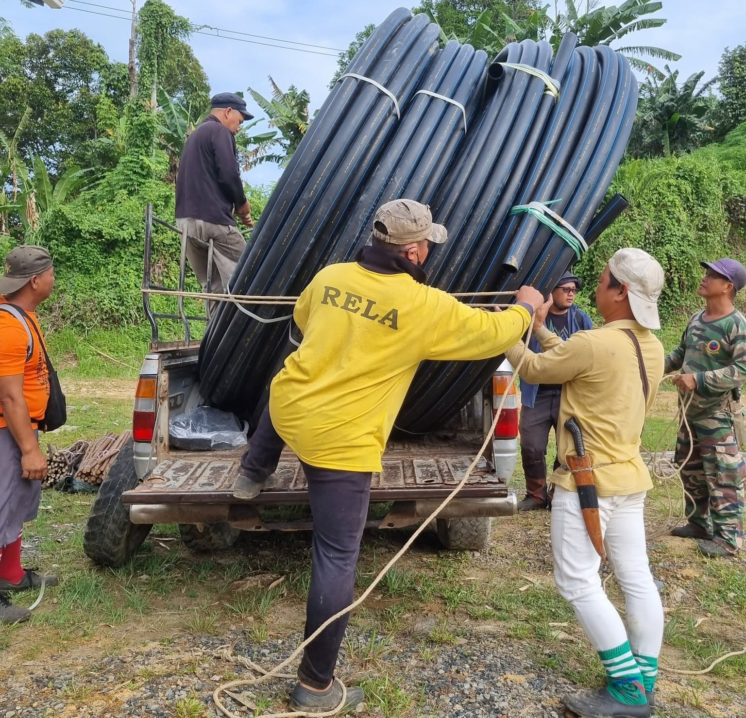 Local villagers and community workers working together to secure large bundles of black HDPE water pipes on a pickup truck during a gravity-fed water supply project in rural Sabah.
