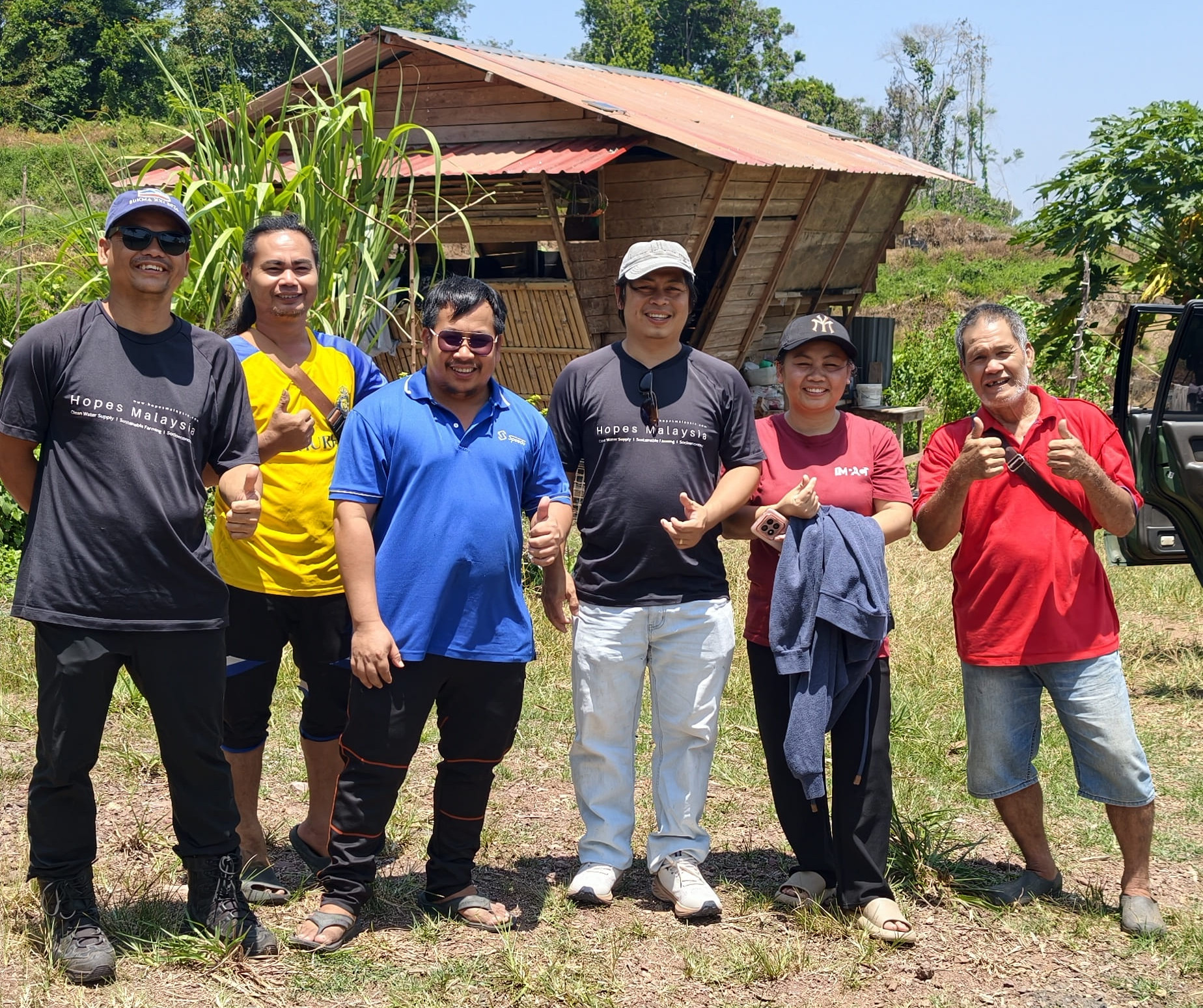 RuralKita field team and local villagers posing for a group photo outdoors in front of a wooden village hut in Kampung Talukung, Kota Belud, Sabah.