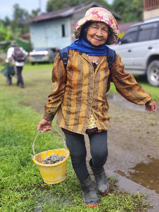 An elderly woman carries a small bucket filled with rocks to support her community’s gravity water project construction.