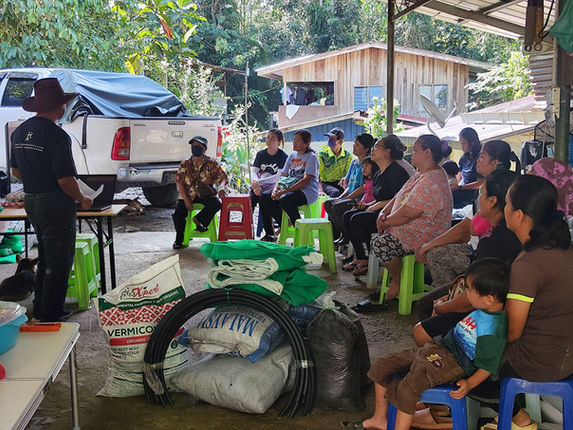 Rural community members seated together during an outdoor village meeting beside a wooden house, with a pickup truck and building materials nearby.