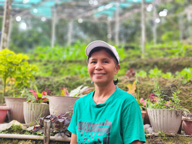 Woman wearing a green shirt and white cap sitting in an outdoor garden with potted plants and greenery behind her.