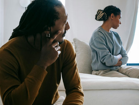 Man in brown sweater looks towards a woman in blue, sitting on a bed with arms crossed, appearing distant. Neutral-colored room, daytime.