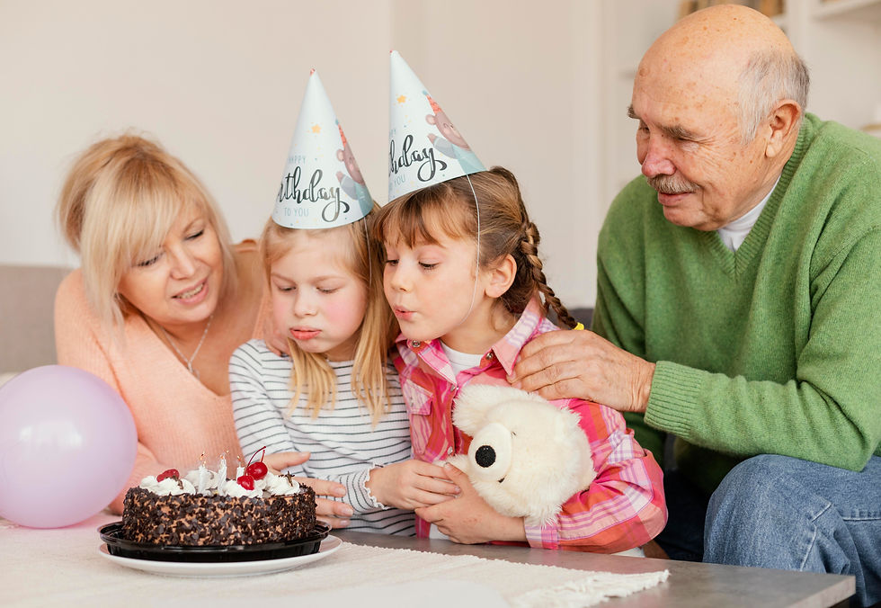 Two kids in party hats blow out candles on a chocolate cake, with an older couple watching. A balloon and stuffed toy are nearby.